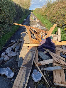 Wood planks, rocks, and debris block a narrow country lane with hedges on both sides.